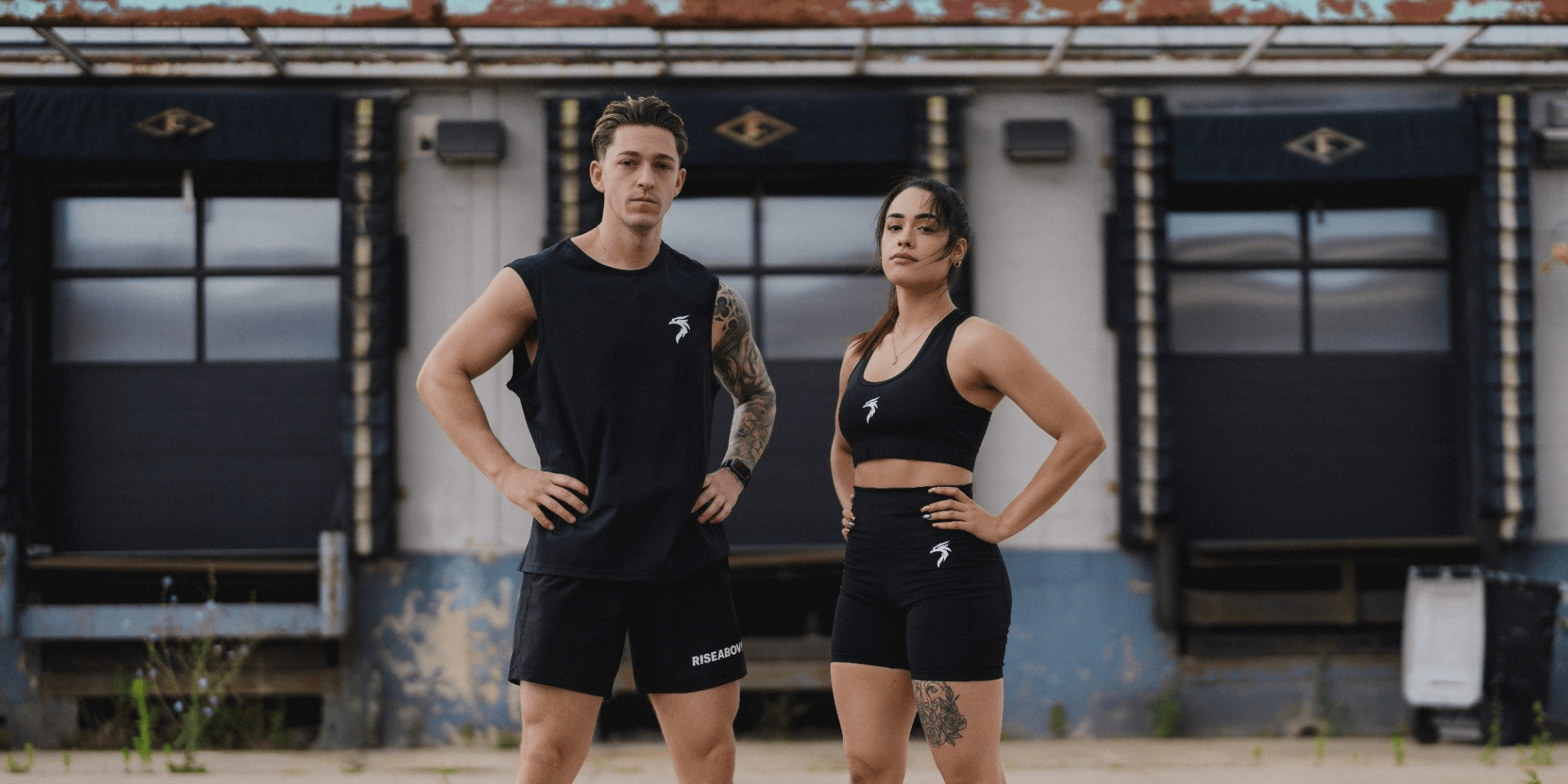 Man and woman wearing Riseabove black athletic activewear outdoors. Men's muscle tank and shorts, women's sports bra and bike shorts.